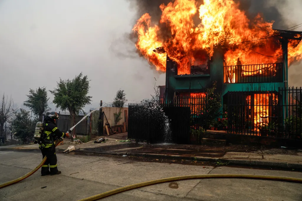 A firefighter tries to extinguish a fire in front of a burning building as fire and smoke rise from a forest fire in the Biobio region where, according to local media, multiple wildfires prompted emergency evacuations, in Concepcion, Chile, January 18, 2026.