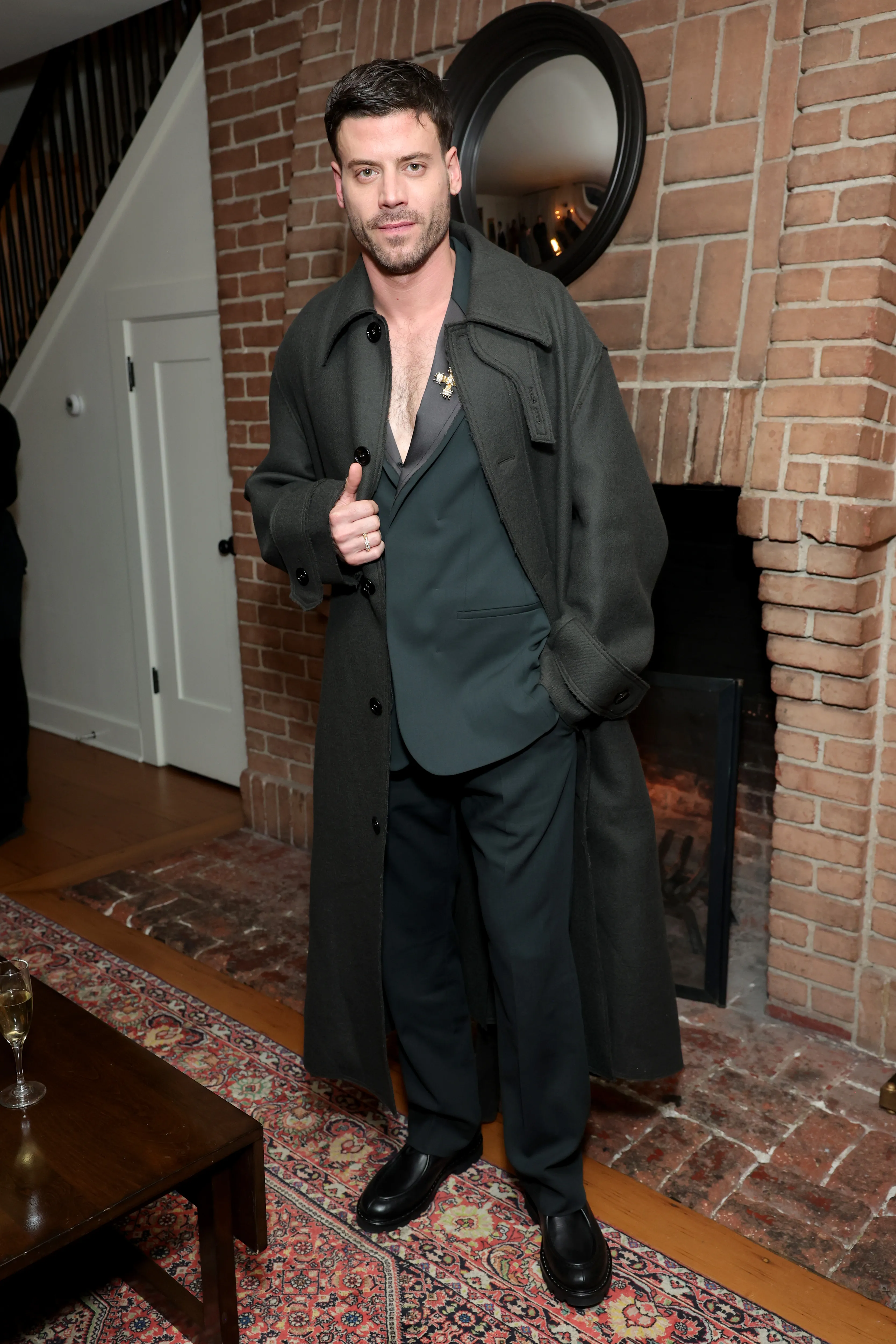 François Arnaud posing in a long dark coat and an unbuttoned suit jacket, with a brick fireplace behind him.