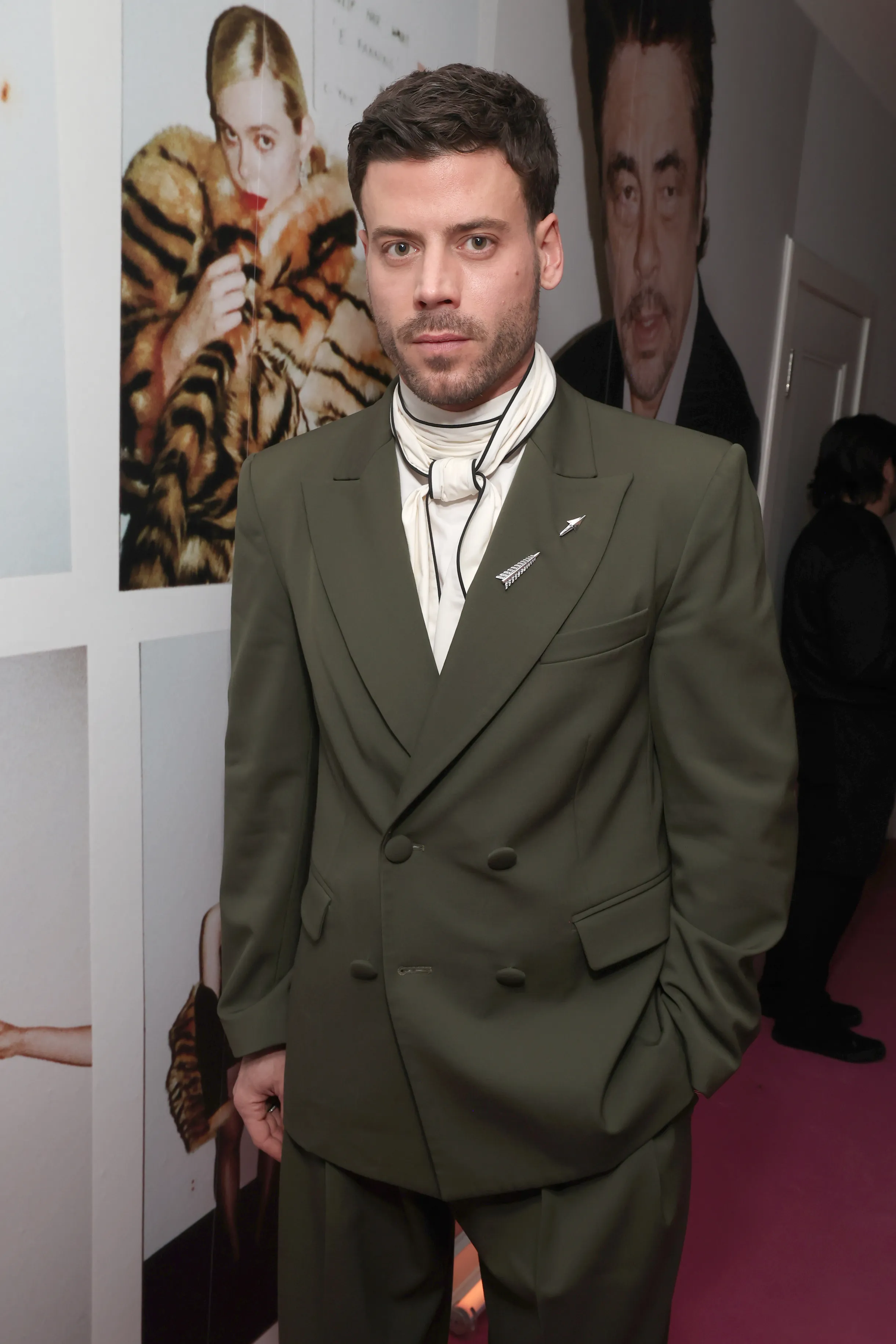 François Arnaud in a dark green double-breasted suit with a cream scarf and arrow-shaped brooches.