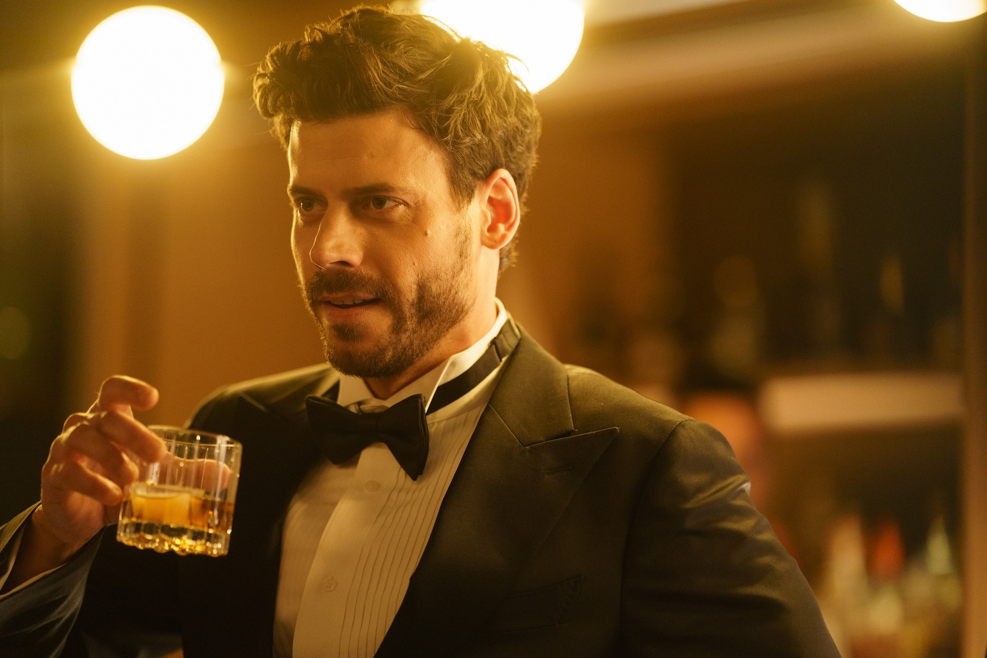 François Arnaud as Scott Hunter, holding a glass of whiskey.