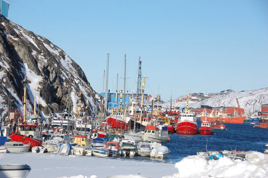 General view of the port in Nuuk with numerous boats, snow-covered hills, and buildings in the background.