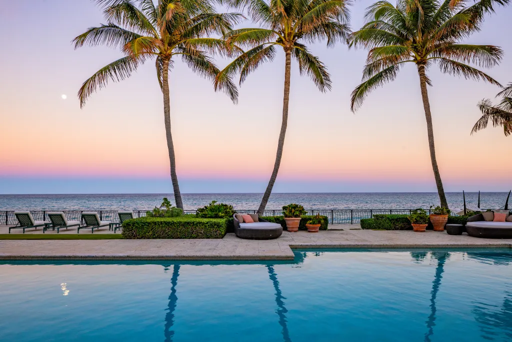 An outdoor pool, lounge chairs, and palm trees in front of the ocean during sunset in Manalapan.
