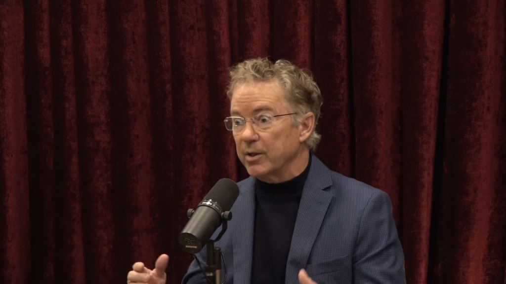 Rand Paul speaking into a microphone with a red curtain background.