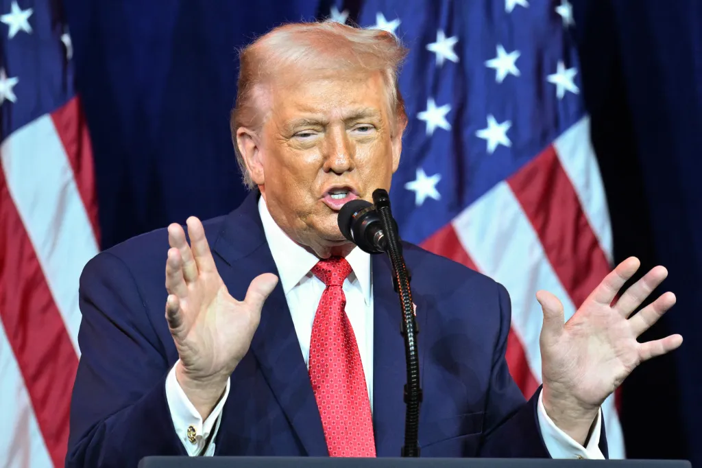 Former US President Donald Trump speaking at a podium with two American flags in the background.