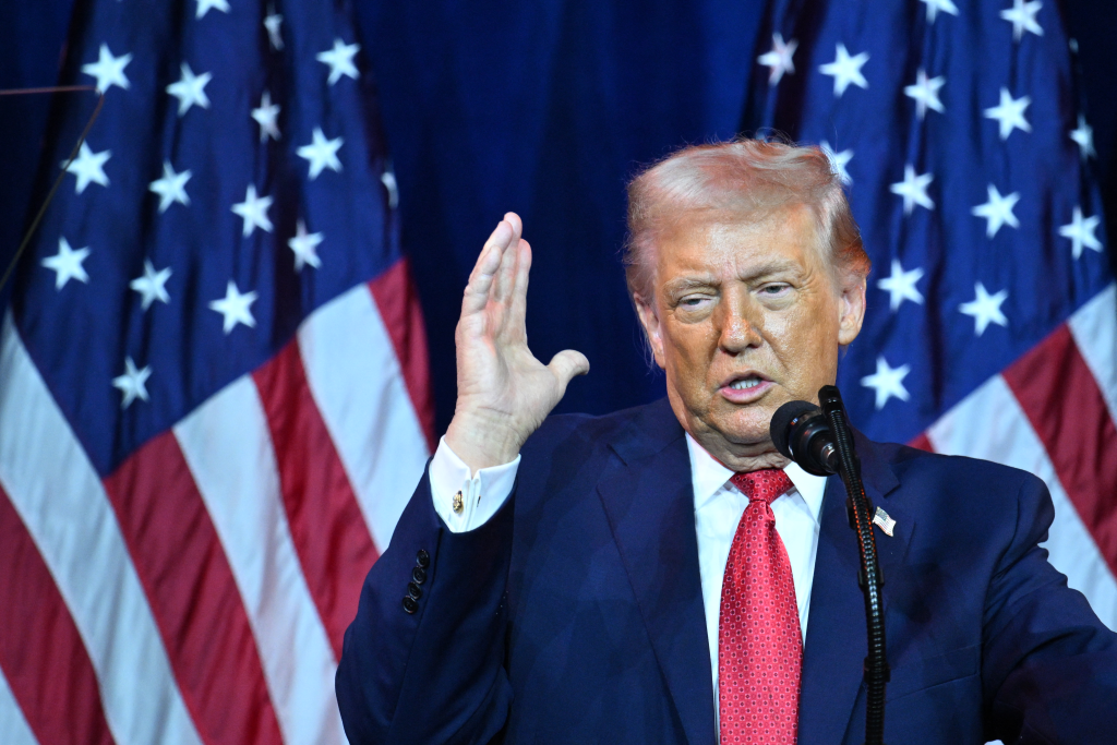 Donald Trump speaking at a podium with a microphone, gesturing with his right hand, in front of American flags.