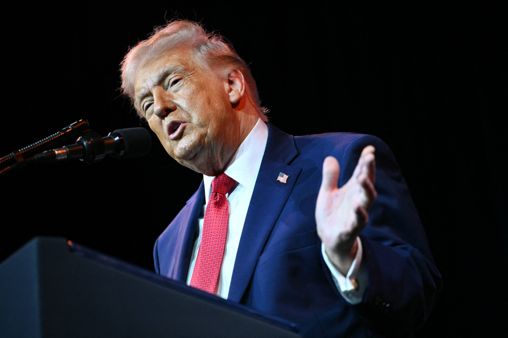 Donald Trump speaking at a podium with a microphone, gesturing with his left hand.