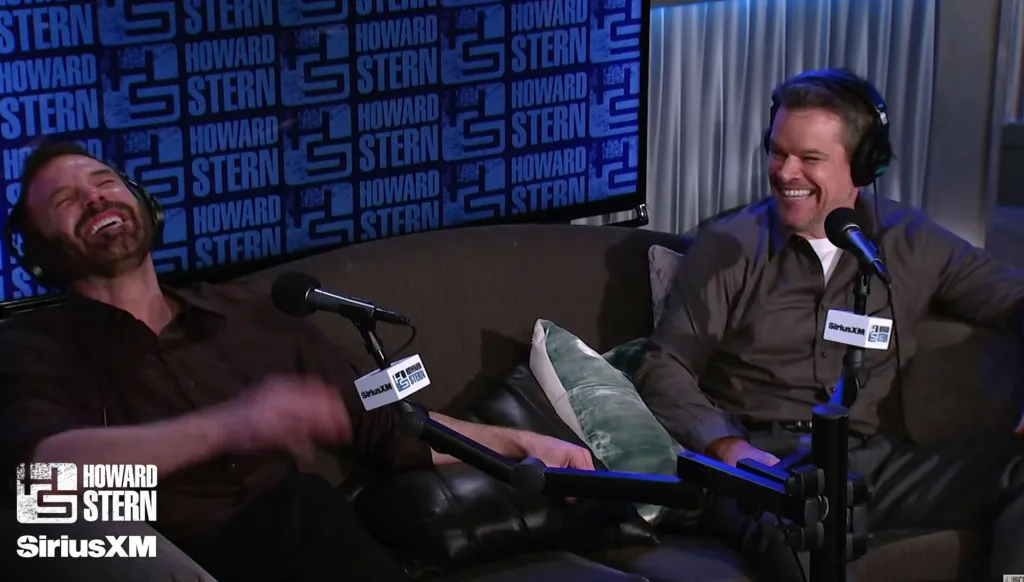 Matt Damon and Ben Affleck laughing on a couch during an interview for Howard Stern on SiriusXM.