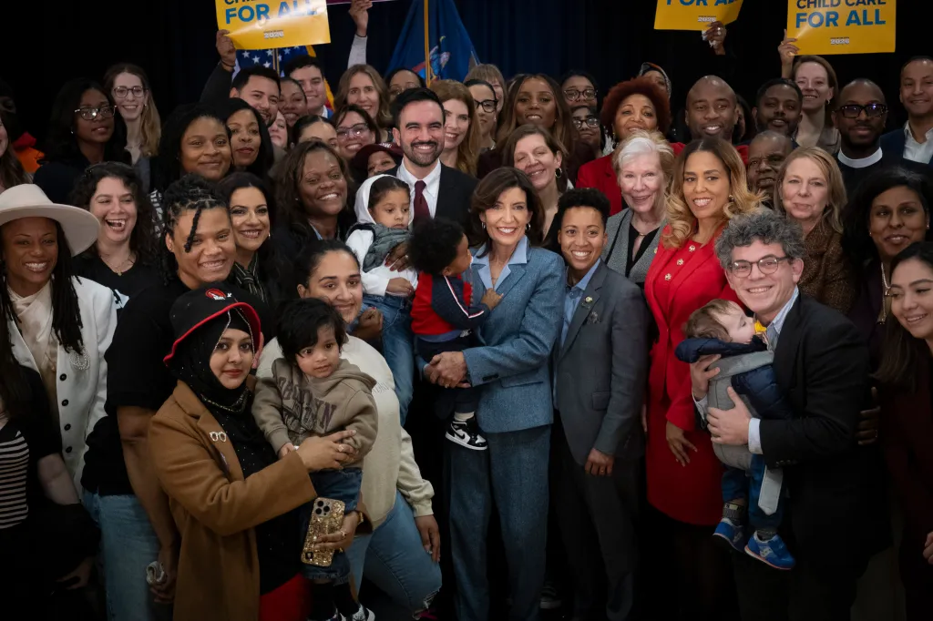 Governor Kathy Hochul and Mayor Mamdani pose with a diverse group of people, including several young children, with signs for 