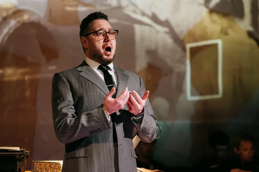 A man in a pinstripe suit and glasses sings with his mouth open, holding his hands up.