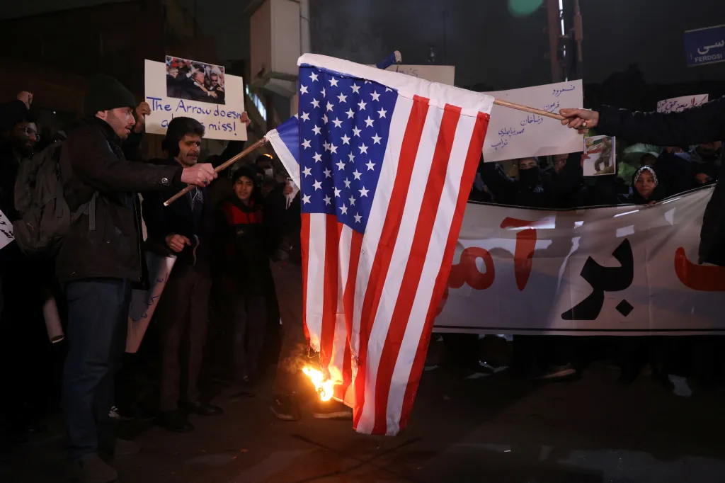 Protesters in Tehran burn a US flag, with signs in Farsi and English visible in the background.