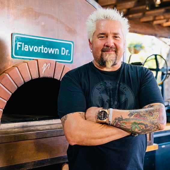 Guy Fieri stands in front of a brick oven with a 