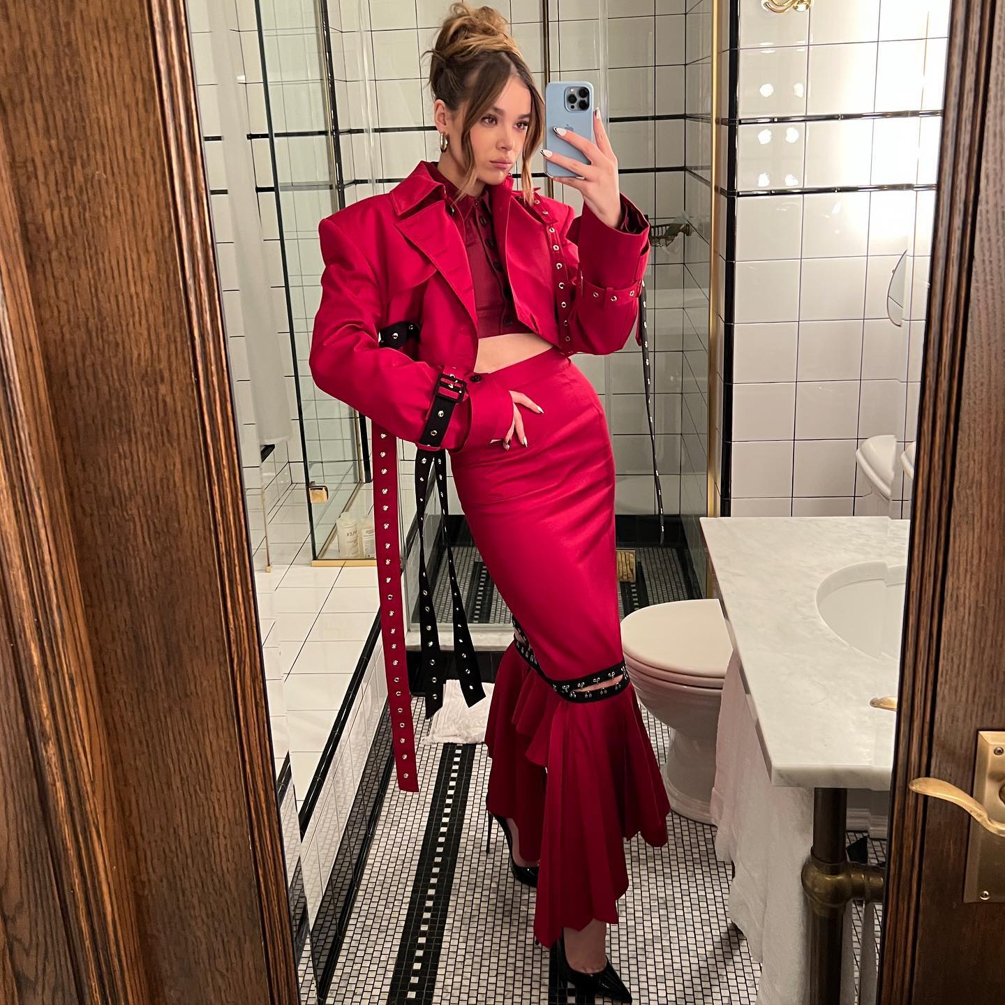 Hailee Steinfeld taking a mirror selfie in a red co-ord skirt and jacket, with black studded details.