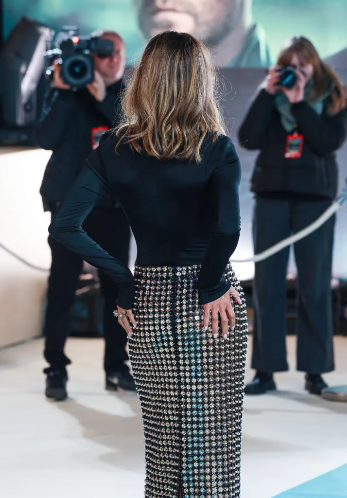 Halle Berry on a red carpet wearing a black long-sleeved top and a dazzling, floor-length skirt made of small, reflective beads, with her hands resting on her hips.