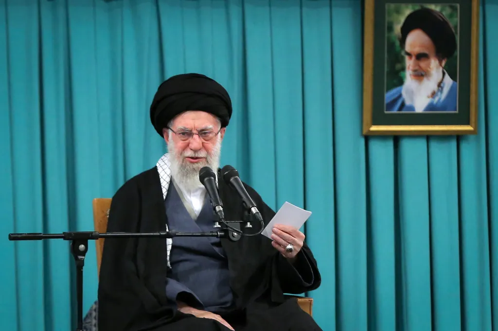 Ayatollah Ali Khamenei addressing a meeting with microphones in front of him and a framed portrait of Ruhollah Khomeini in the background.