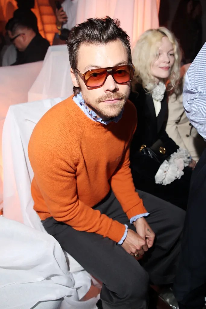 Harry Styles in an orange sweater and large sunglasses, sitting at the Valentino Pavillon des Folies Show during Paris Fashion Week.