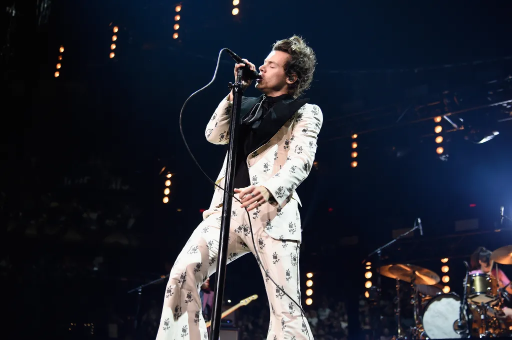 Harry Styles performing onstage in a white and black floral suit, singing into a microphone.