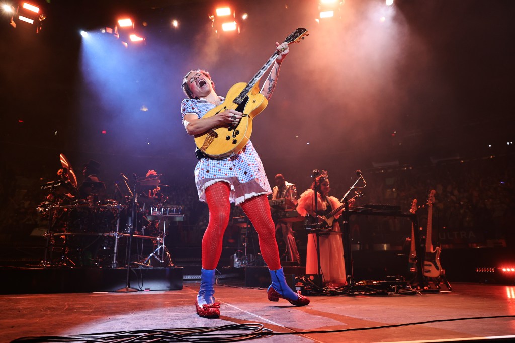 Harry Styles performing onstage in a Dorothy costume, holding a yellow guitar above his head and singing.