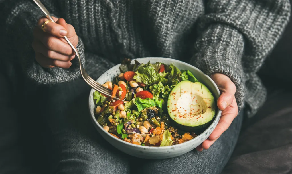 A person in a gray sweater and jeans eating a Buddha bowl with a fork.