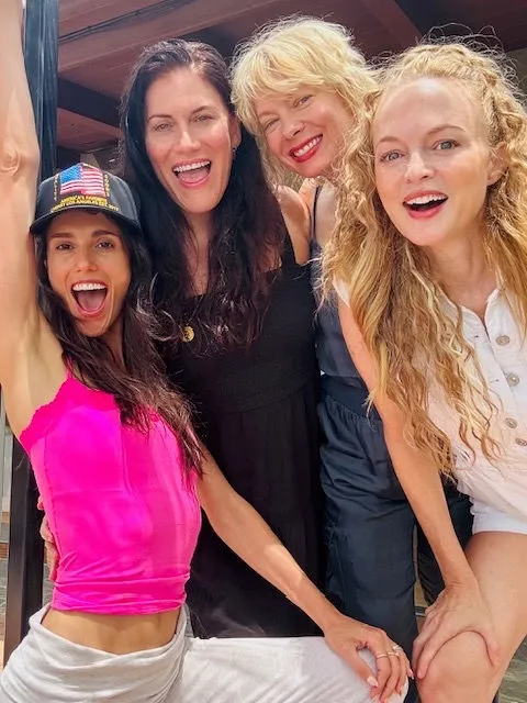 Heather Graham, in a white romper, and three women smiling at the camera.