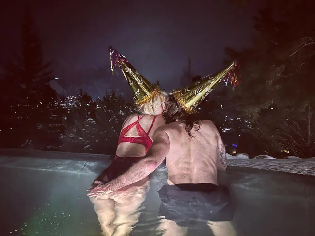 Heidi Klum and Tom Kaulitz wearing party hats and kissing in a hot tub outdoors at night, surrounded by snow-covered trees.
