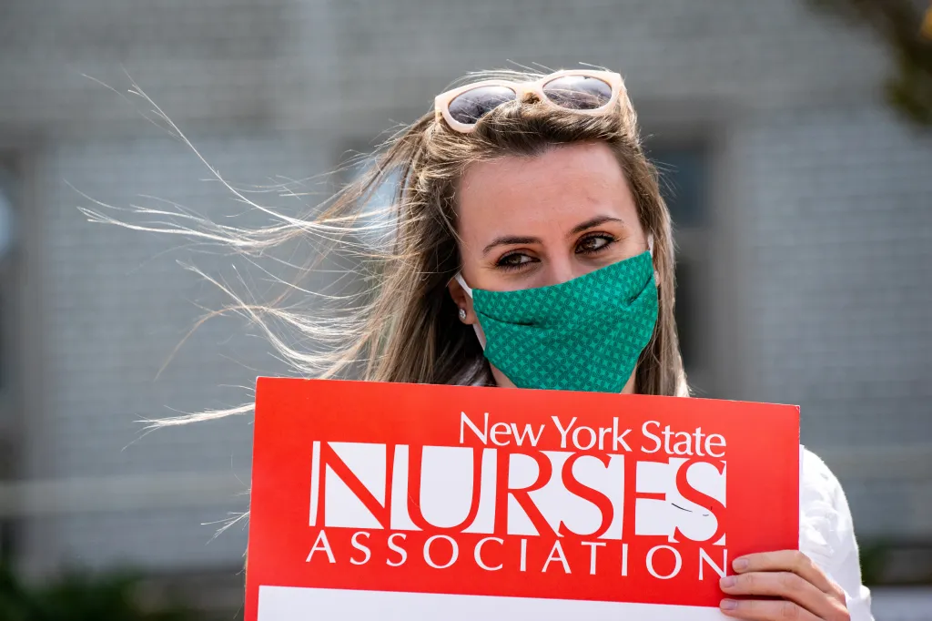 A nurse in a green patterned face mask and sunglasses holds a red sign that reads 