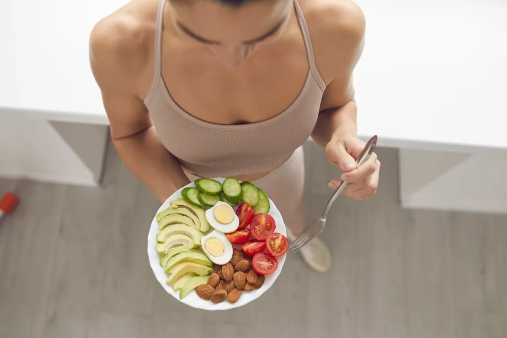 Woman holding a plate with a healthy meal of almonds, egg, sliced avocados, tomatoes, and cucumbers.
