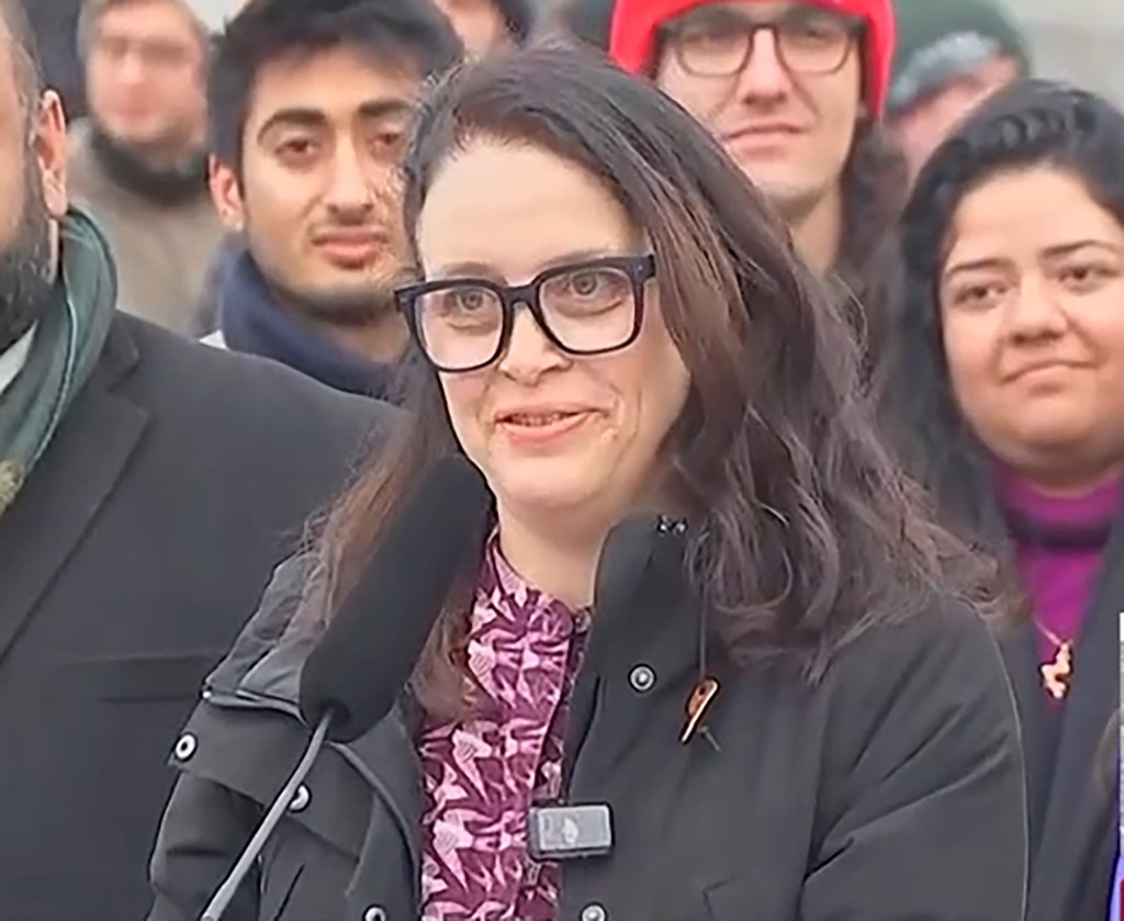 Tascha Van Auken at a press conference, wearing glasses and a black coat over a patterned purple shirt.