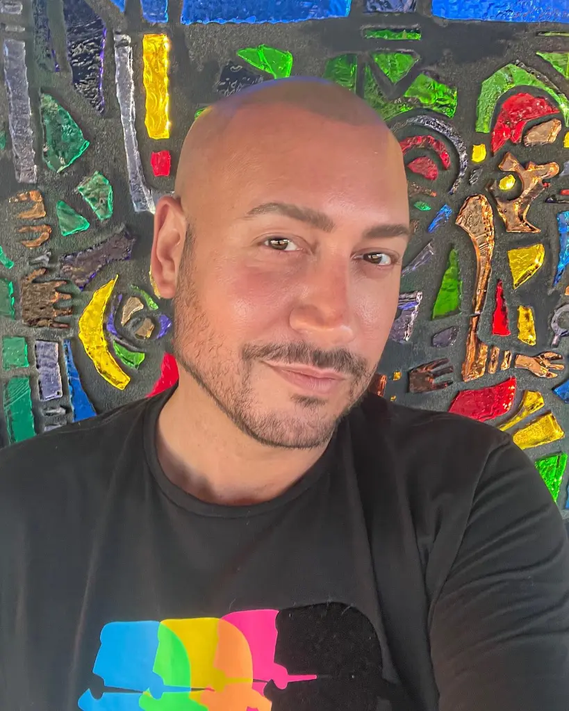 A man in a black shirt with a colorful graphic on the chest stands in front of a colorful stained glass background.