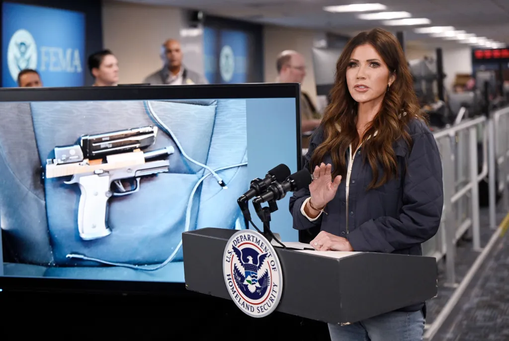 Homeland Security Secretary Kristi Noem holds a news conference with a screen showing a handgun to her left.