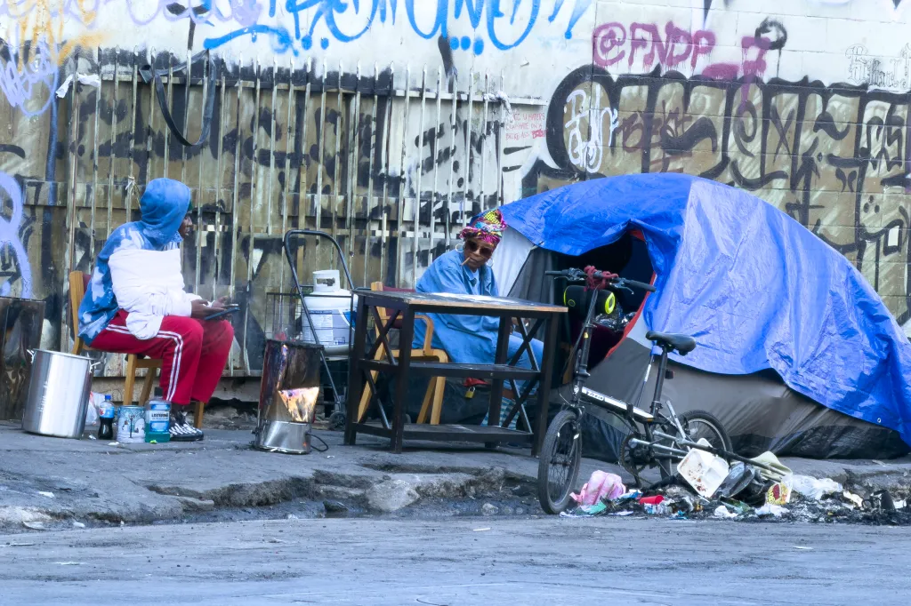 Two homeless people on Skid Row sitting next to tents and belongings on a street.
