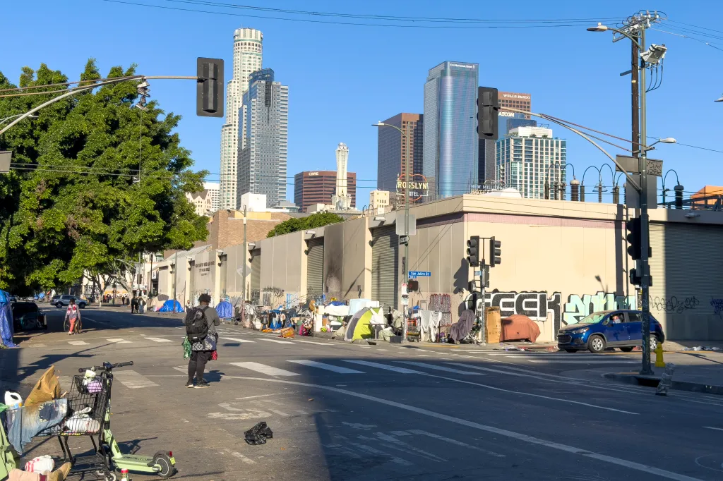 Homeless encampments line a Los Angeles street with tall buildings in the background.