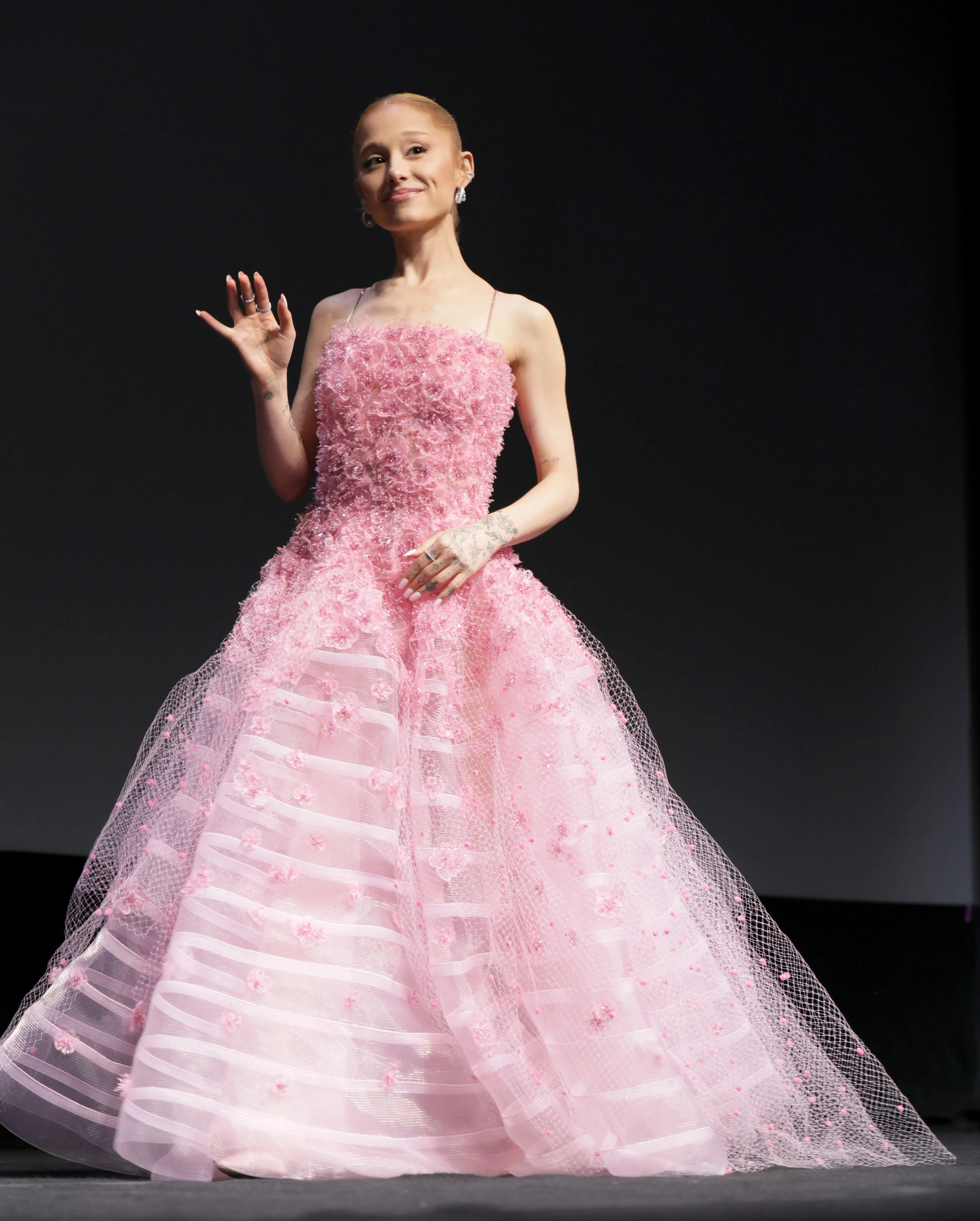 Ariana Grande wearing a pink gown and earrings, standing onstage, smiling.