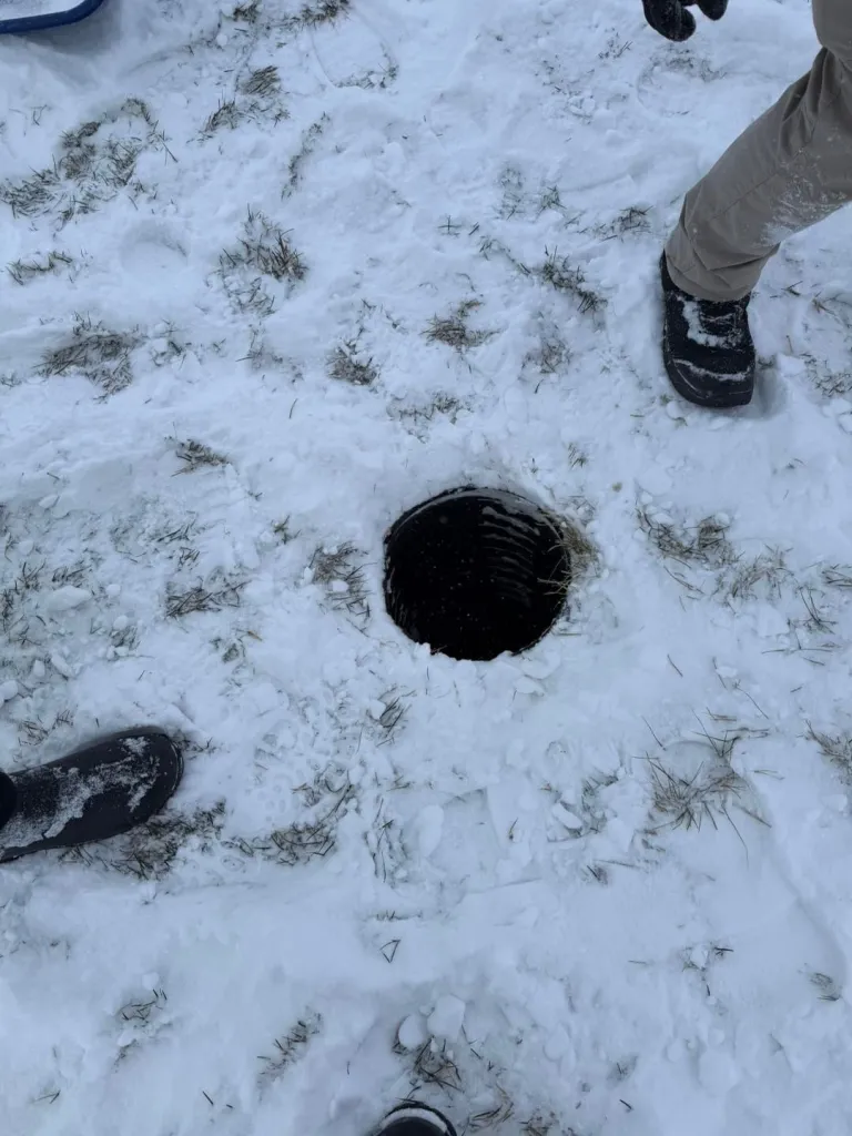 A large, dark hole in the snow next to a person's feet.
