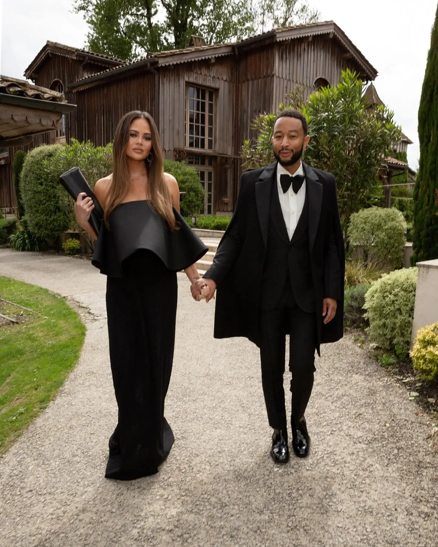 Chrissy Teigen and John Legend holding hands, walking on a gravel path in front of a rustic wooden building.