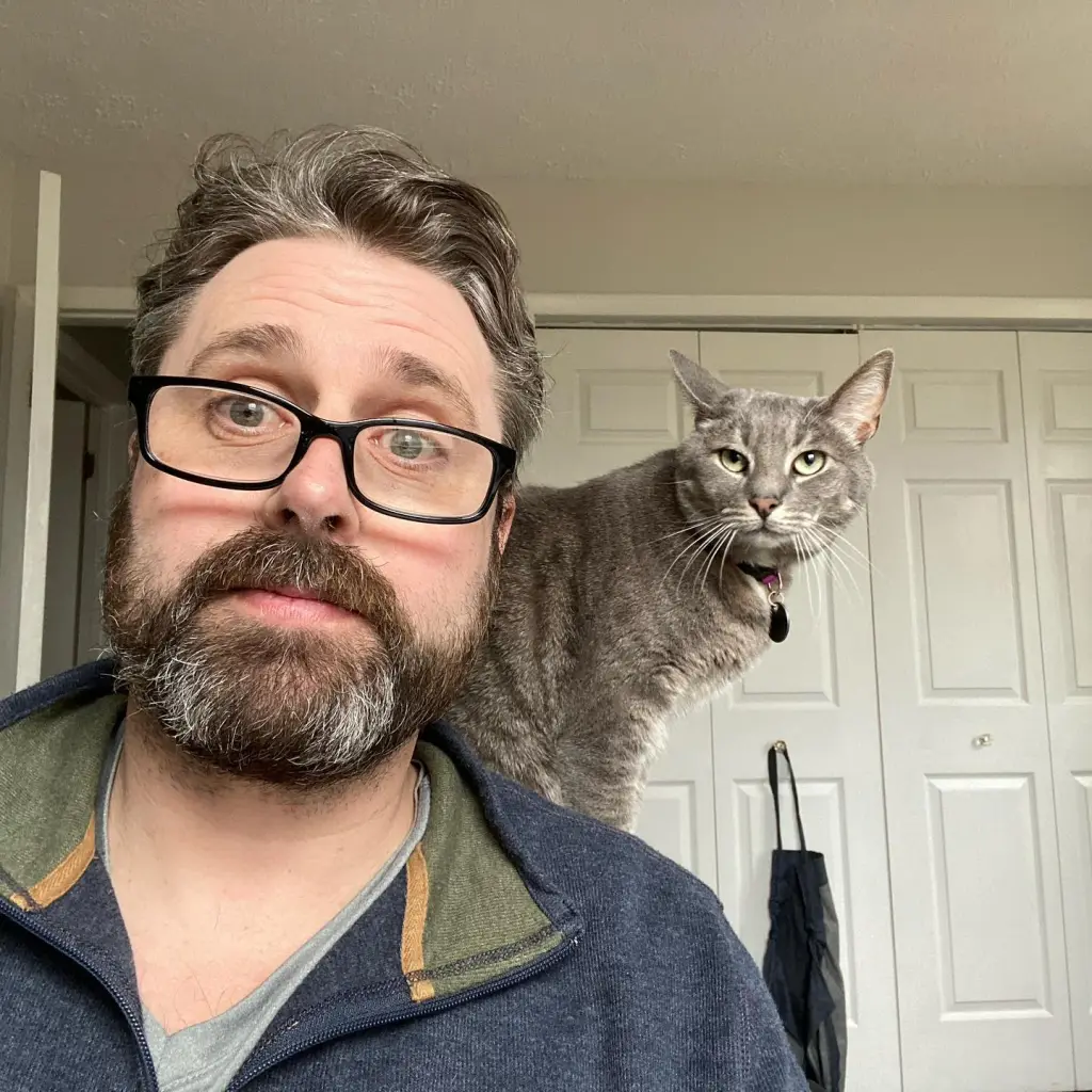 A man with a beard and glasses looks wide-eyed at the camera with a gray cat on his shoulder.