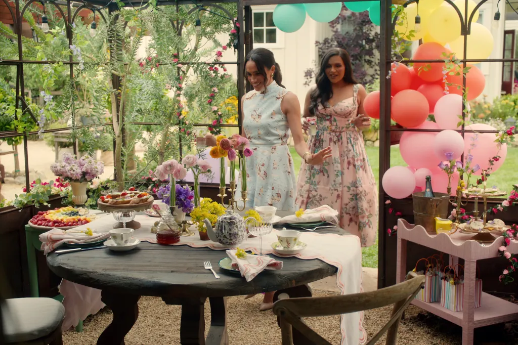 Meghan, Duchess of Sussex,in a floral dress, and Mindy Kaling, in a pink floral dress, at a party table with food, flowers, and balloons.