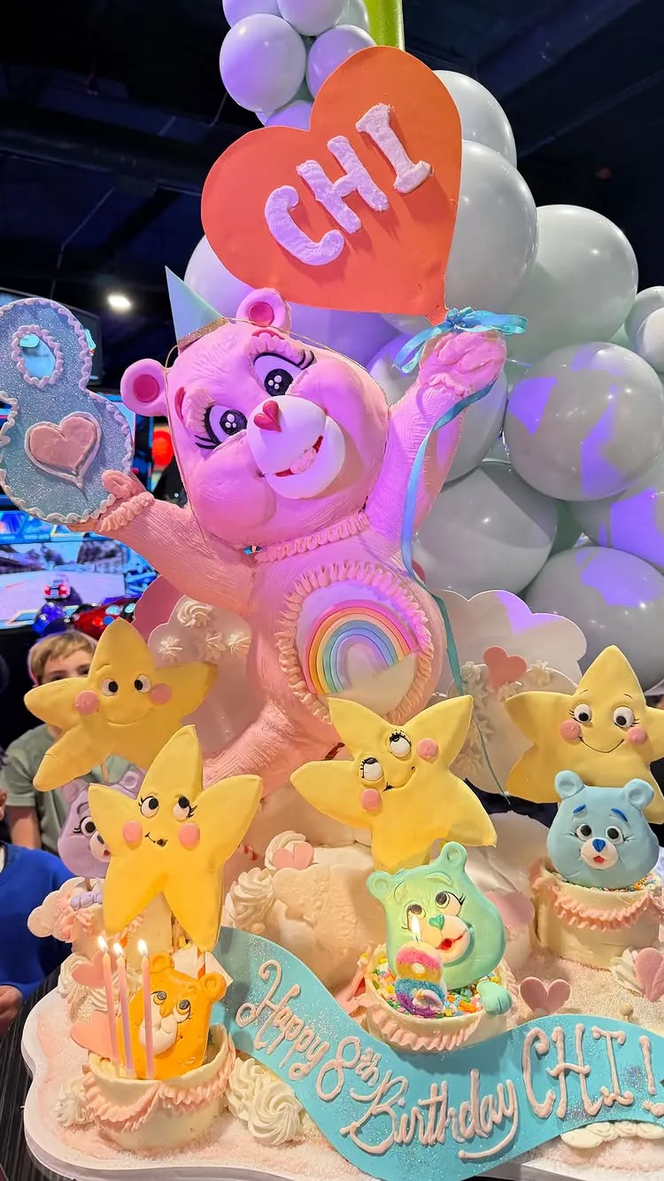 An elaborate, multi-tiered birthday cake in a Care Bear theme, featuring a large pink Care Bear, yellow stars, and a banner reading 