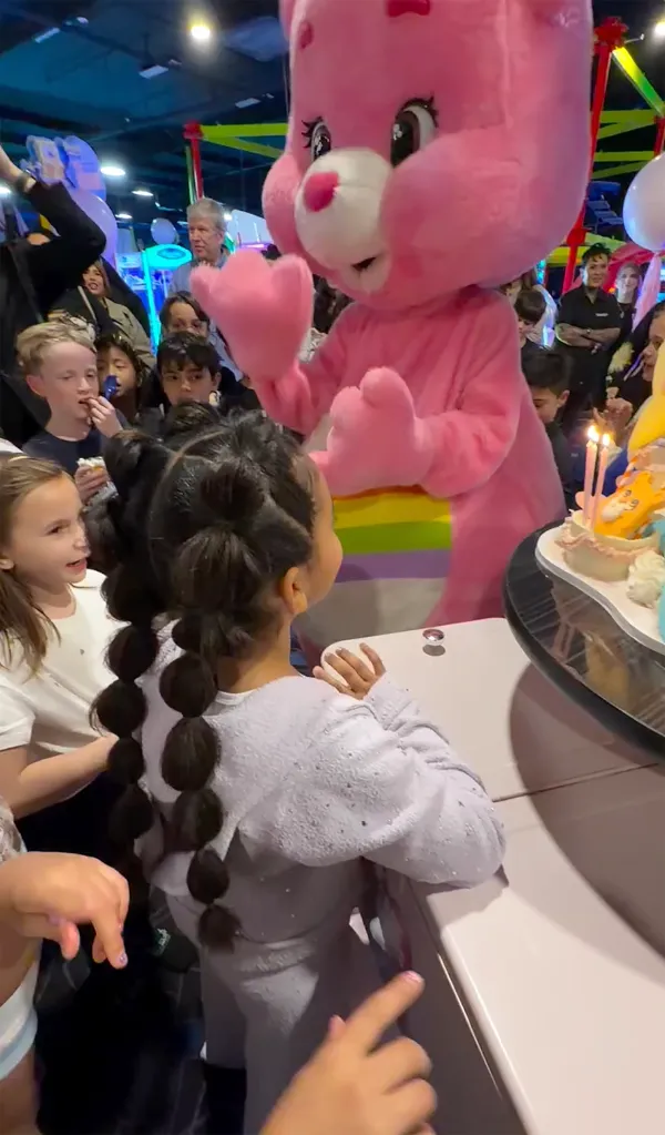 Chicago West in a grey sweater with bubble braids watching a person in a pink Care Bear costume with a rainbow stripe.