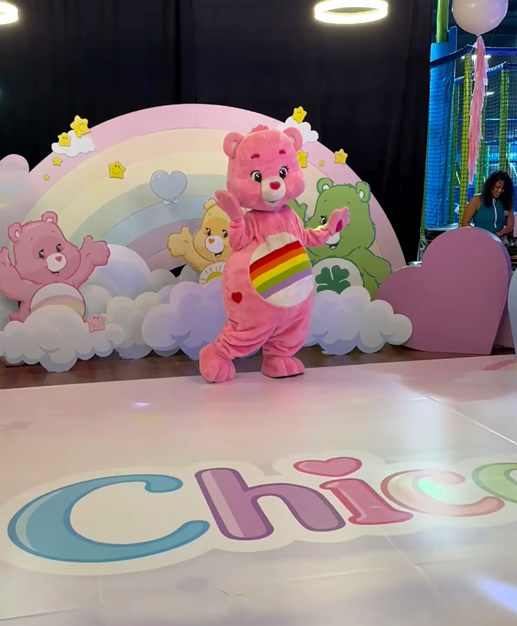 A person in a pink Care Bear costume with a rainbow belly stands on a floor with the name 