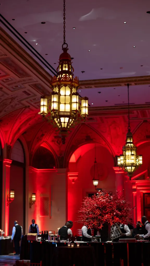 Ballroom decorated with red lights and ornate chandeliers, with servers preparing tables.