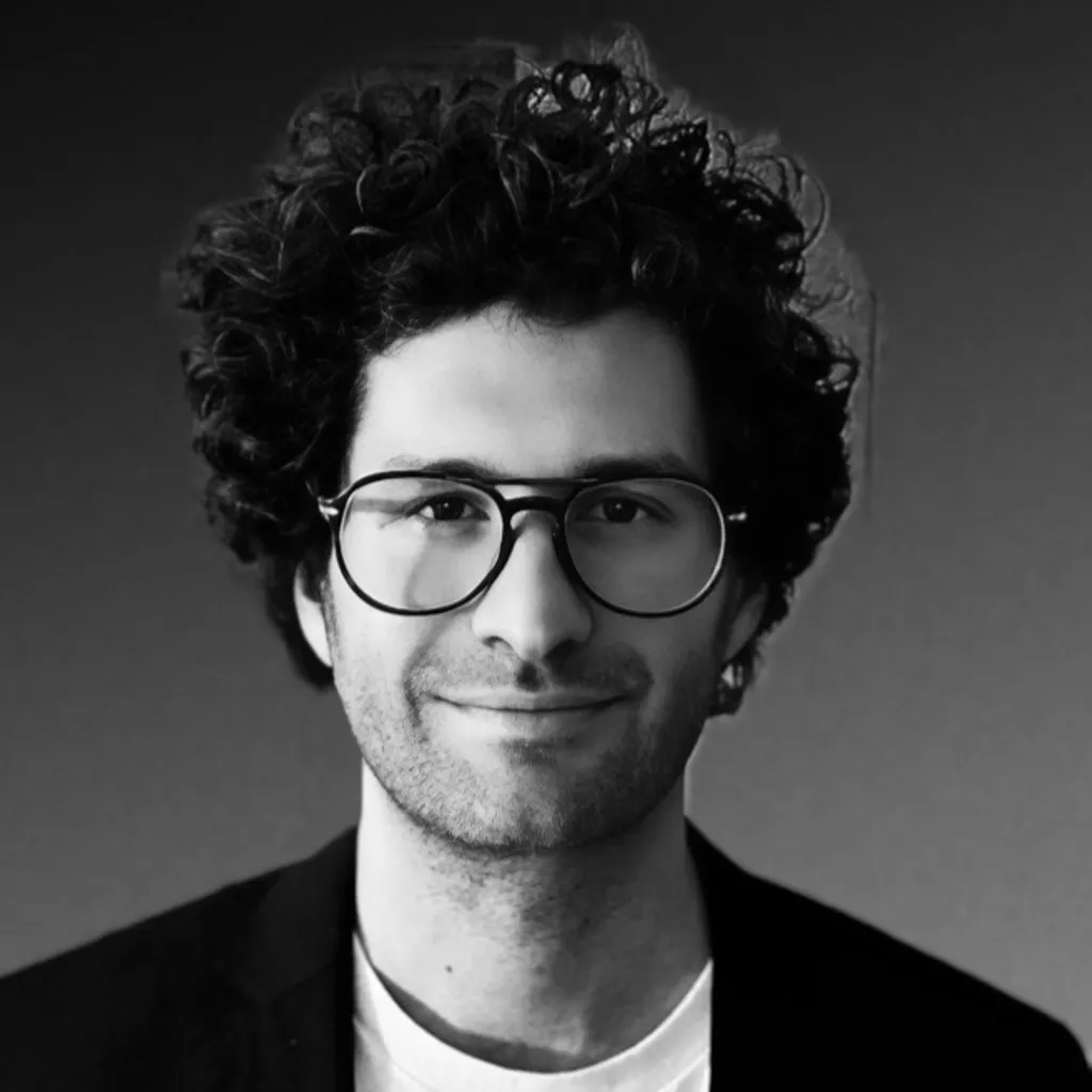 Headshot of Austin Sosa, a man with curly hair and glasses, wearing a white shirt and dark jacket, smiling slightly.