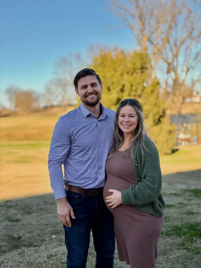 Stephen Wissmann and Jana Duggar, pregnant with their first child, smiling at the camera outdoors.