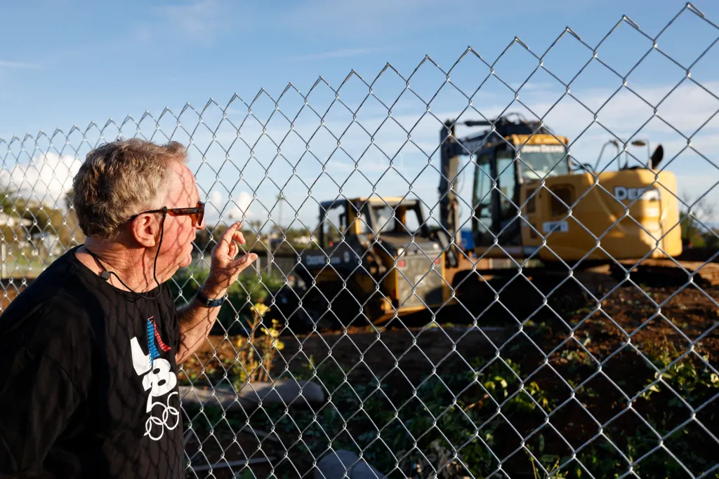Bud King looks at the remains of his home from behind a chain-link fence, with construction vehicles in the background.