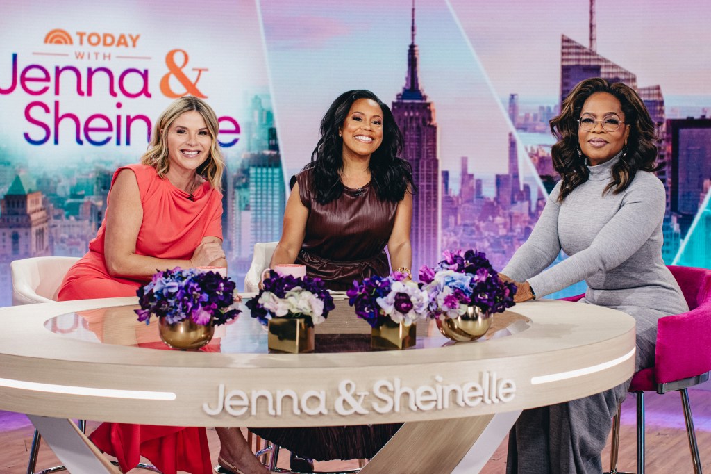 From left, Jenna Bush Hager,in a tangerine dress, Sheinelle Jones, in a burgundy dress, and Oprah Winfrey, in a red dress, seated at a light wood table with a sign reading 