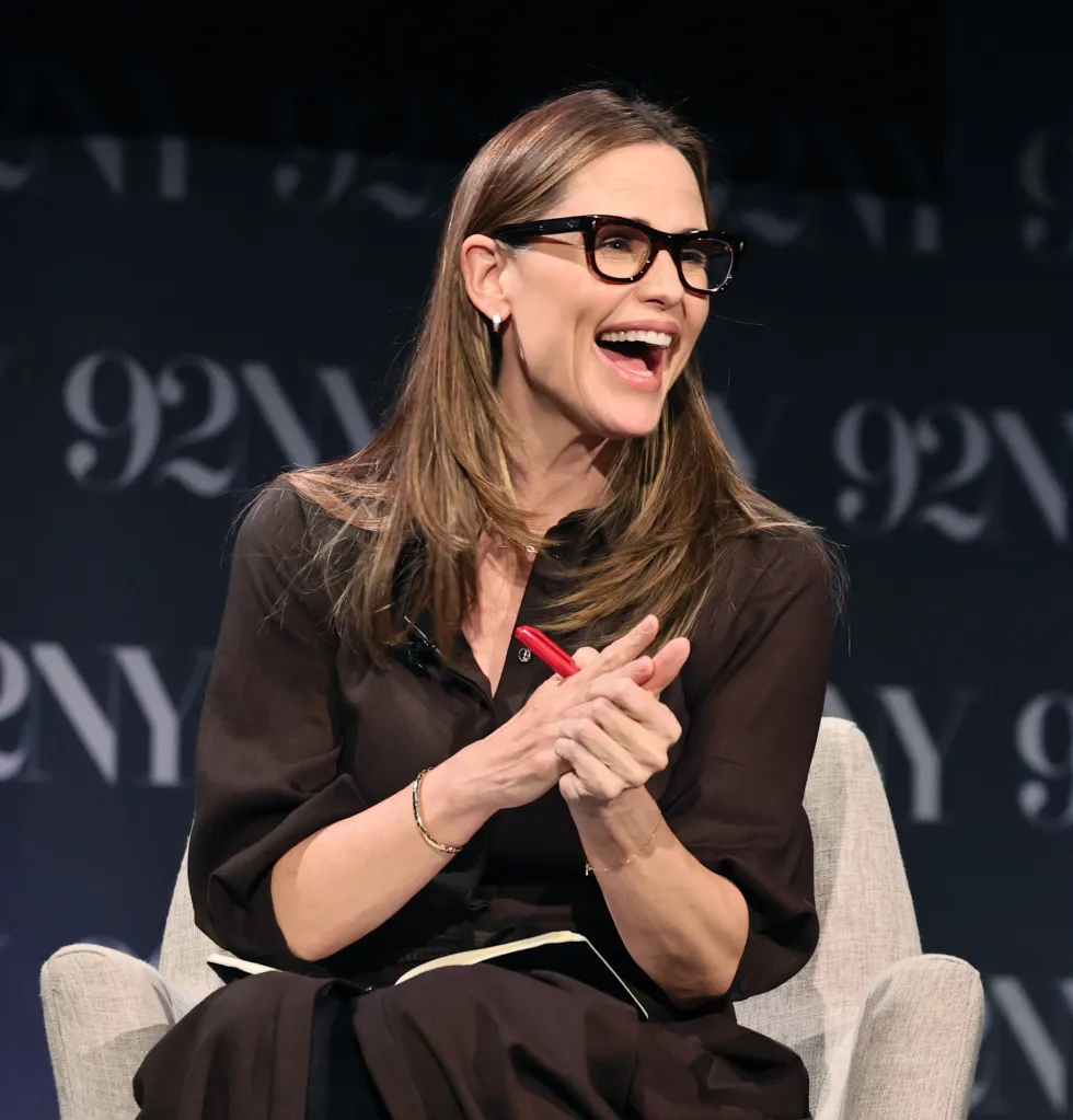 Jennifer Garner laughing while wearing dark-rimmed glasses and a dark brown shirt, with her hands clasped.