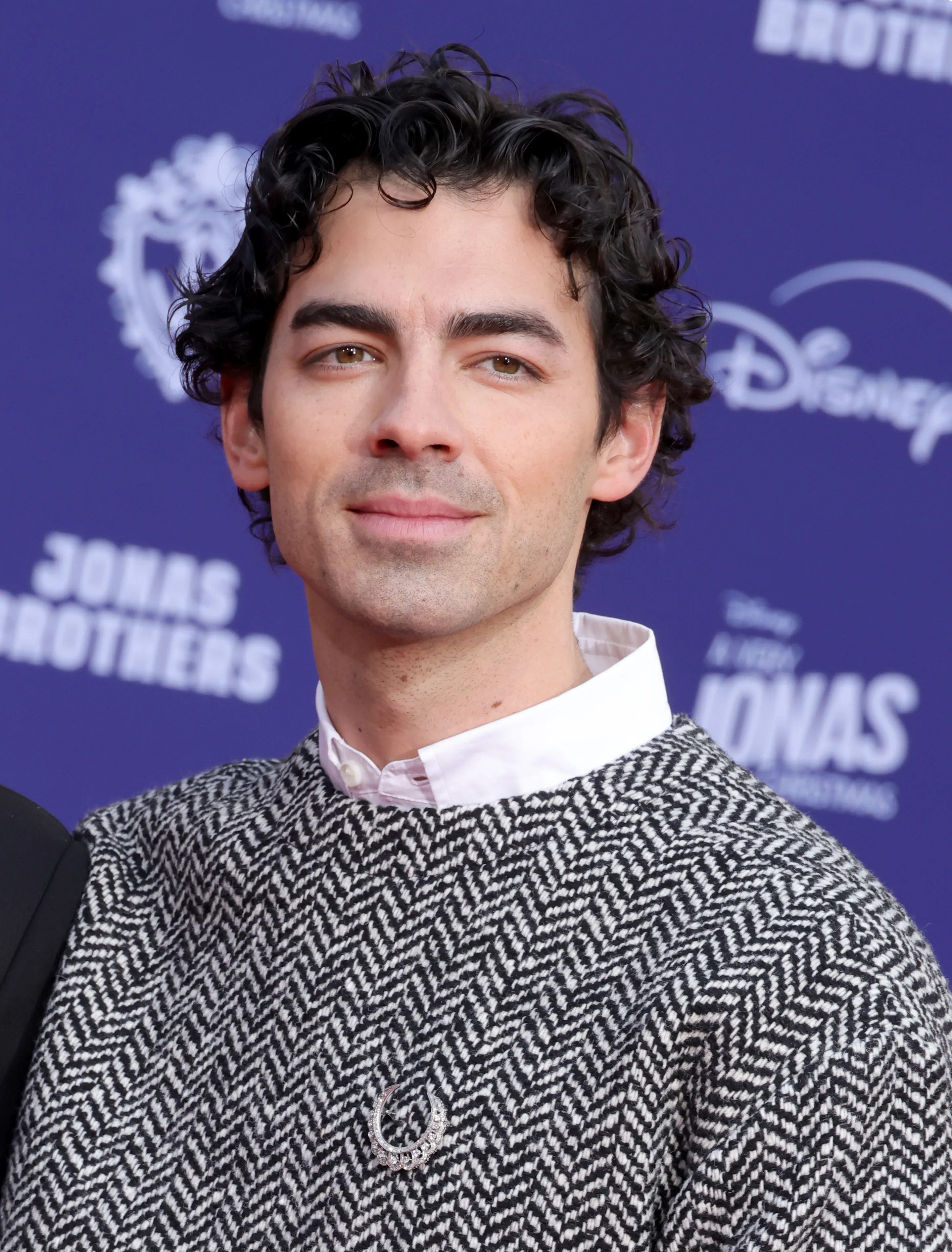 Joe Jonas with dark curly hair wearing a black and white patterned jacket over a white shirt.
