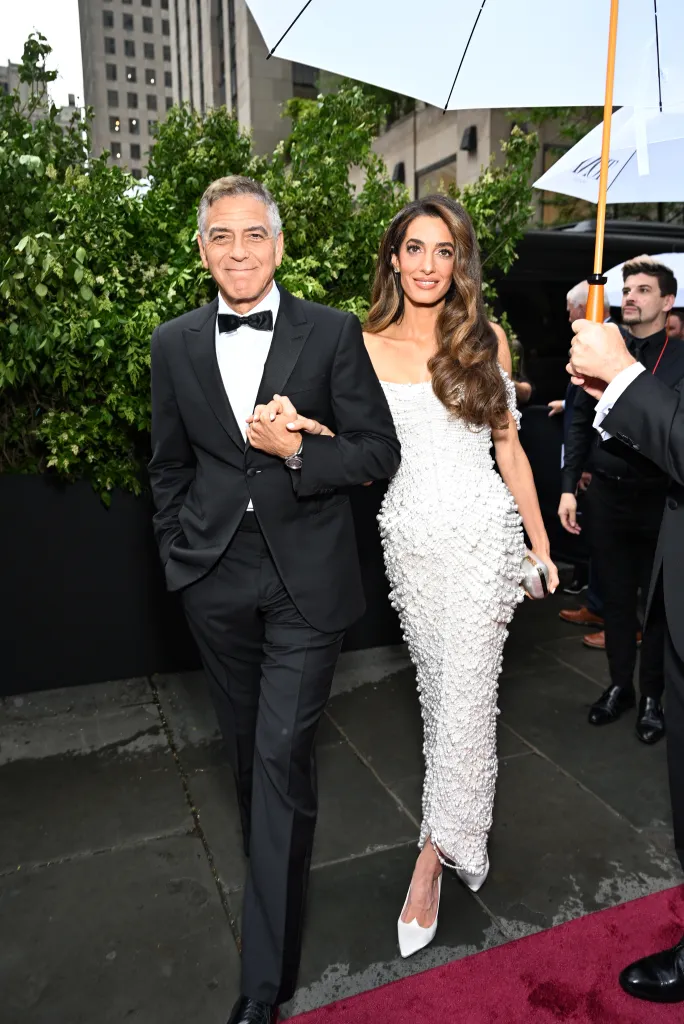 George Clooney in a tuxedo and Amal Clooney in a white, beaded dress holding hands under an umbrella on a red carpet.