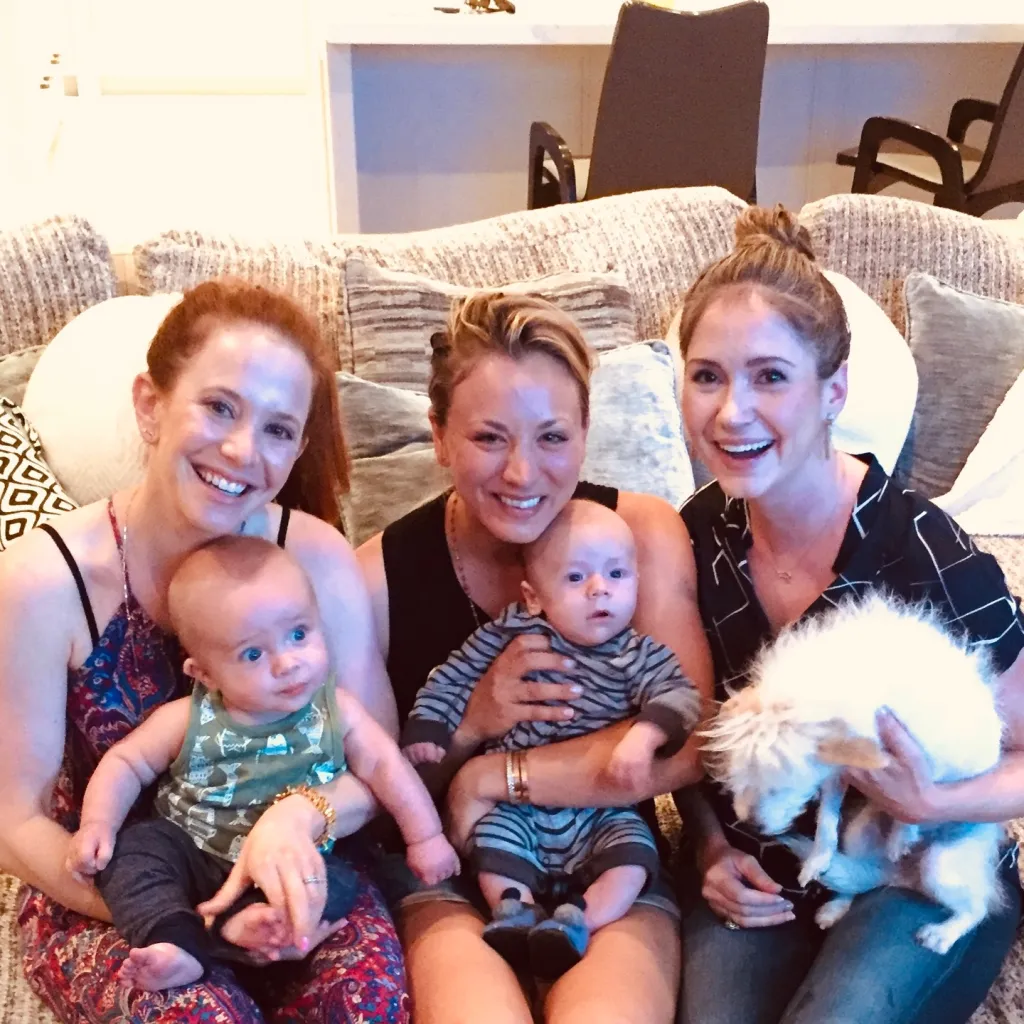 Kaley Cuoco and two other women sit on a sofa holding babies and a fluffy white dog.