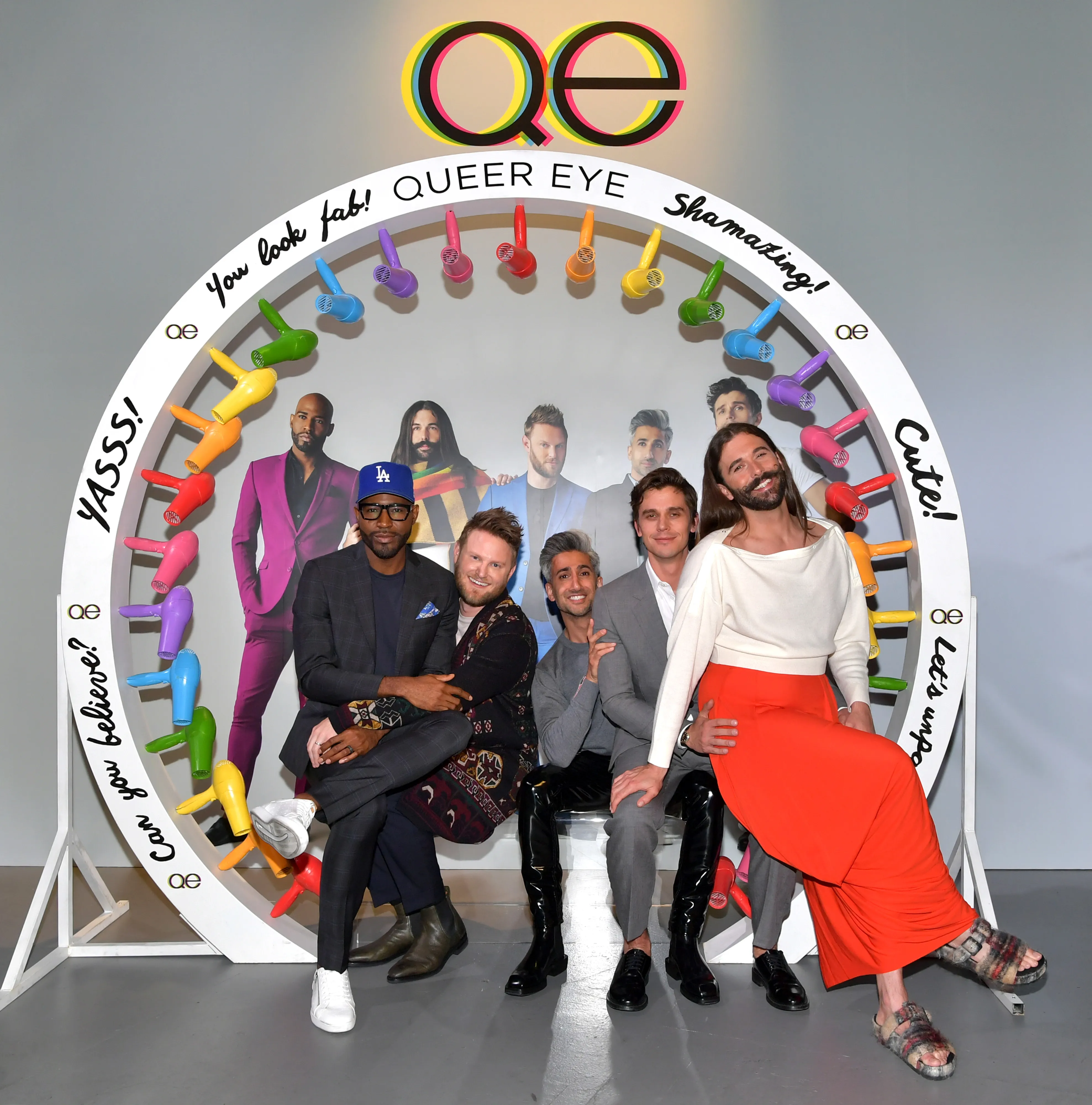 Cast members of Queer Eye pose in front of a white circular backdrop featuring colorful hair dryers and phrases.