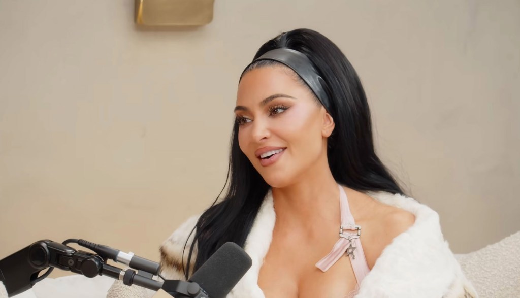 Kim Kardashian smiling while wearing a black headband, white fur coat, and light pink top, with a microphone in front of her.